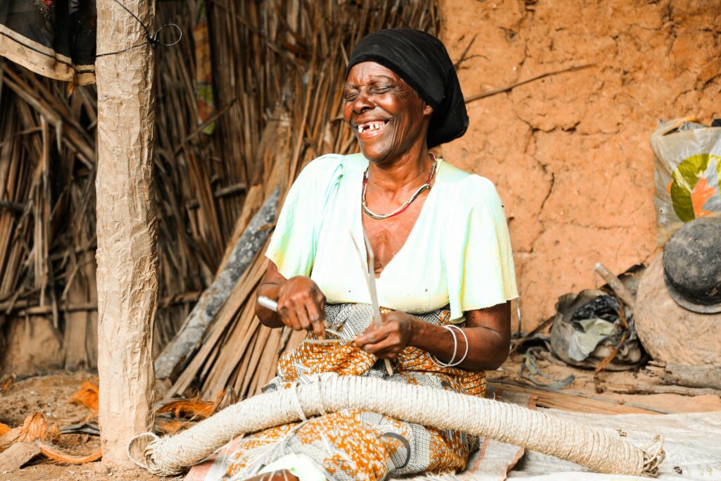 pexels-photo-3307279-3307279 An African woman smiles as she weaves in a rural Tanzanian setting, showcasing traditional craftsmanship.